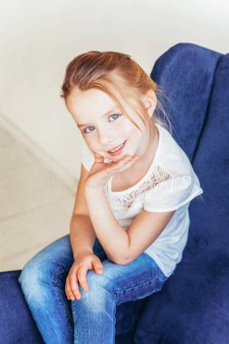 Kot pantolonlu, beyaz tişörtlü, modern, rahat, mavi koltukta oturan, evdeki beyaz, parlak oturma odasında dinlenen tatlı küçük kız. Çocukluk çocukları gençlik konsepti.