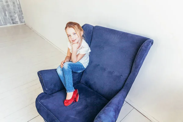 ジーンズの甘い女の子と白のTシャツは、屋内の自宅で白い明るいリビングルームでリラックス現代的な居心地の良い青い椅子に座っています。子供の頃の若者のリラックスコンセプト. — ストック写真