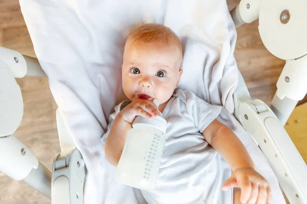 Aranyos újszülött lány tejet iszik üvegből, és fehér háttérrel nézi a kamerát. A csecsemő csecsemő szopogatja a tejet, miközben otthon fekszik az etetőszéken. Anyaság boldog gyermek — Stock Fotó