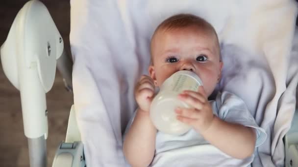 ボトルからミルクを飲んで、白い背景のカメラを見てかわいい新生児の女の子。乳児は自宅の給餌椅子に横になってミルクの栄養を食べて吸う。母親の幸せな子供 — ストック動画