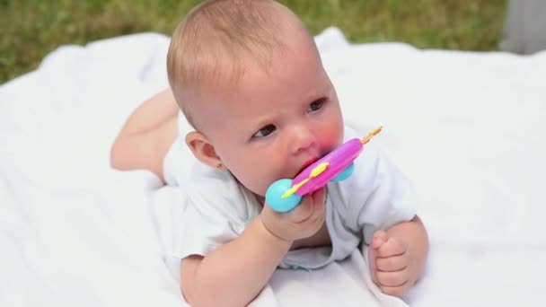 Little newborn girl lying on tummy chewing rattle on blanket, summer day outdoor. Infant having fun outdoors. Infant baby child resting playing learning to crawl. Motherhood teething babyhood concept — Stock Video