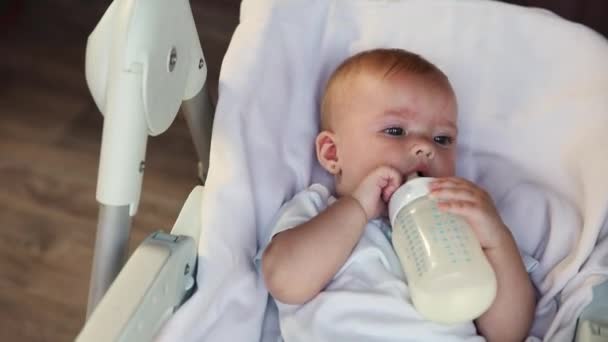 सुंदर लहान नवजात मुलगी बाटलीतून दूध पिते आणि पांढरा पार्श्वभूमीवर कॅमेरा पाहते. अर्भक बाळ घरी आहार खुर्चीवर खाली पडलेले दूध पोषण खात आहे. मातृत्व आनंदी मुलगा — स्टॉक व्हिडिओ