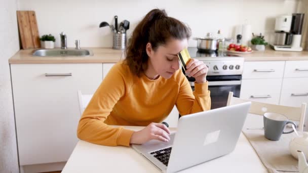 Femme faisant du shopping en ligne et payant par carte de crédit en or. Jeune fille assise avec ordinateur portable achetant sur Internet entrez les détails de carte de crédit sur fond de cuisine intérieure. Boutique en ligne e-commerce concept. — Video