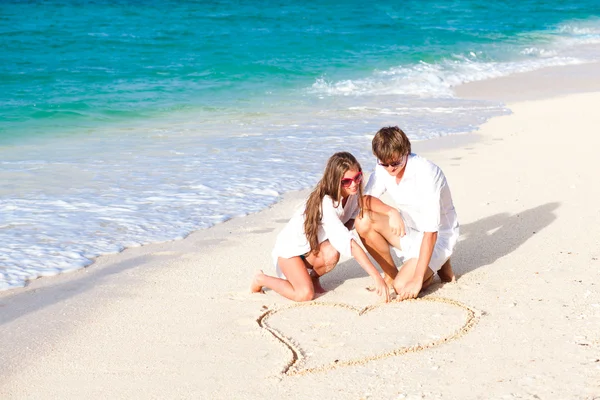 Joven pareja feliz dibujando corazón en la playa tropical. luna de miel —  Fotos de Stock