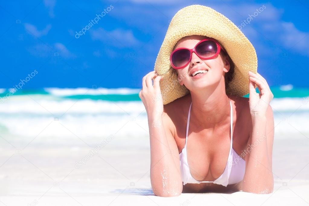 Front view of beautiful young woman in sunglasses and straw hat lying on beach enjoying the sun