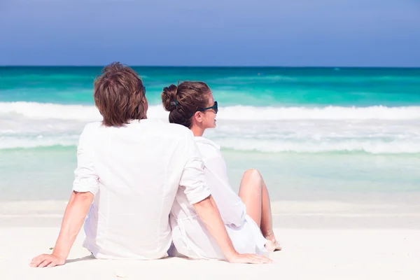Felice giovane coppia in abiti bianchi in occhiali da sole seduto sulla spiaggia — Foto Stock