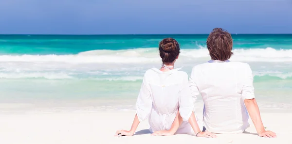 Indietro vista felice giovane coppia in abiti bianchi in occhiali da sole seduto sulla spiaggia — Foto Stock