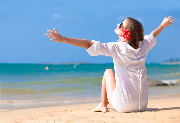 Vista posterior joven mujer de pelo largo en camisa blanca y gafas de sol con flor en el pelo sentado en la playa —  Fotos de Stock