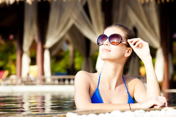Krásná mladá žena v sluneční brýle s květinou ve vlasech s úsměvem v luxusní bazén — Stock fotografie