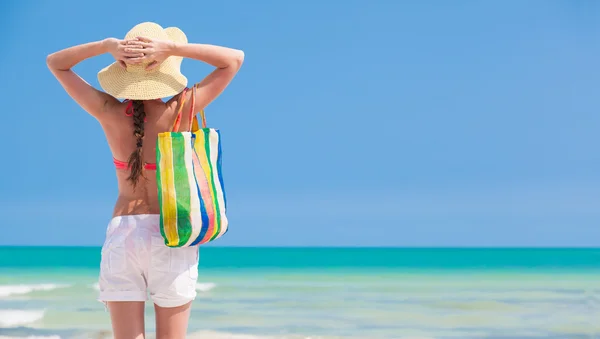 Kvinna i bikini och halm hatt med strandväska stående på stranden. bakifrån — Stockfoto