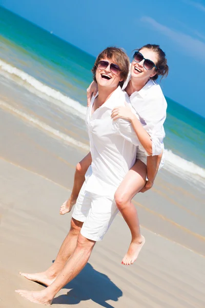 Feliz pareja en gafas de sol y ropa blanca en vacaciones piggybacking alegre en la playa — Foto de Stock