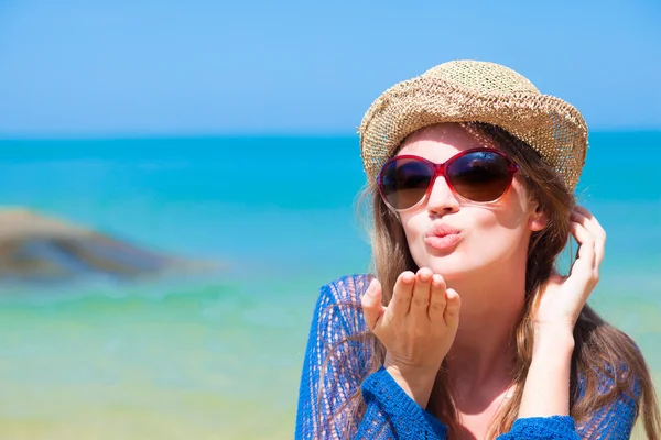 Portrait of young woman in sunglasses and straw hat blowing an air kiss on beach — Stock Photo, Image