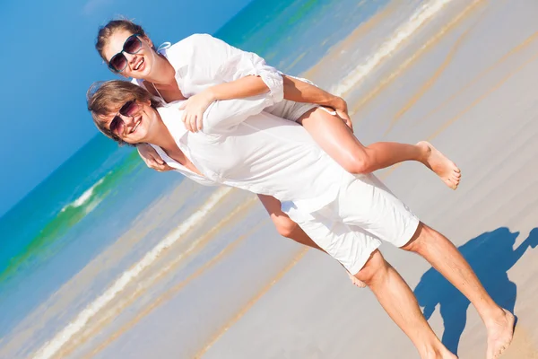Coppia felice in occhiali da sole e vestiti bianchi in vacanza a cavalluccio allegro sulla spiaggia — Foto Stock