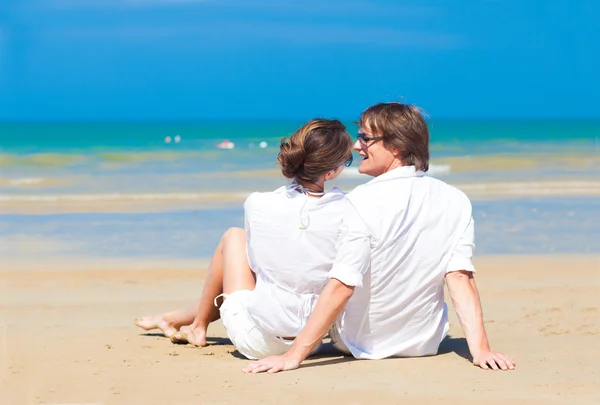 Щаслива молода пара в білому одязі в сонцезахисних окулярах сидить на пляжі — стокове фото