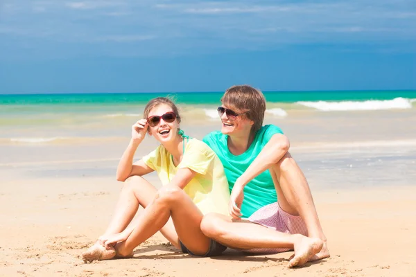 Retrato de feliz pareja joven en gafas de sol en ropa brillante sentado en la playa tropical —  Fotos de Stock