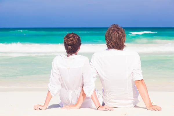 Visão traseira do feliz casal jovem lua de mel em roupas brancas sentado na praia — Fotografia de Stock