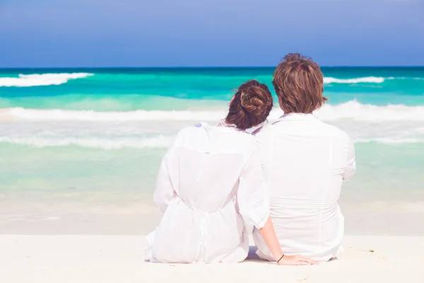 Happy young honeymoon couple in white clothes sitting on beach — Stock Photo, Image