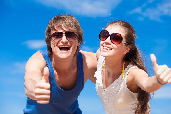 Portrait of happy young couple in sunglasses having fun on tropical beach. Thumbs up