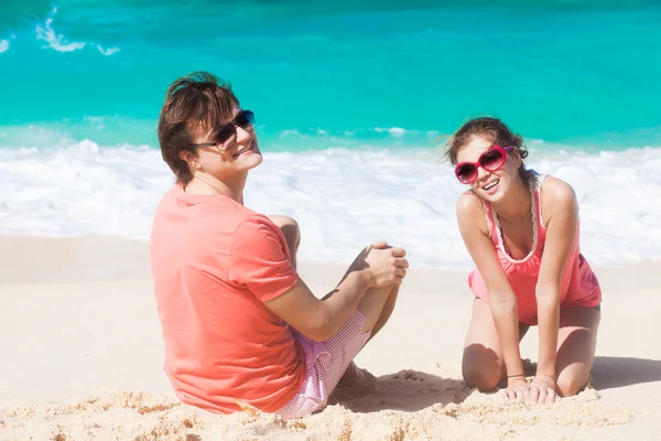 Mladý šťastný Kavkazský pár v sluneční brýle sedí na tropické pláži — Stock fotografie