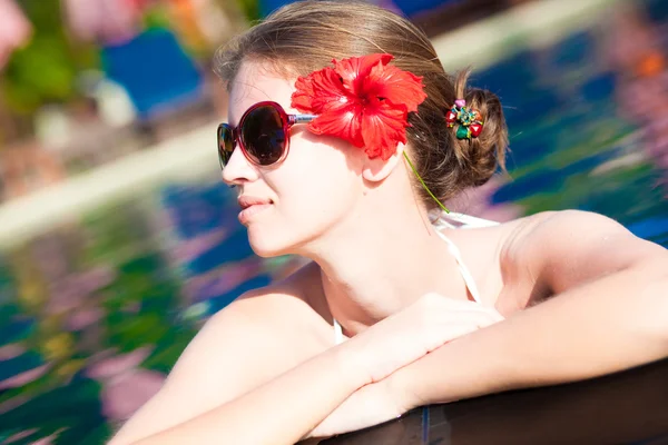 Belle jeune femme en lunettes de soleil avec fleur dans les cheveux souriant dans la piscine de luxe — Photo