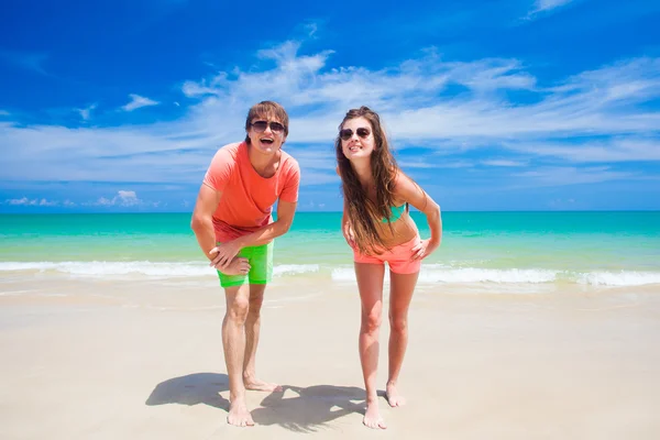 Retrato de jovem casal feliz em roupas brilhantes e óculos de sol se divertindo na praia tropical — Fotografia de Stock