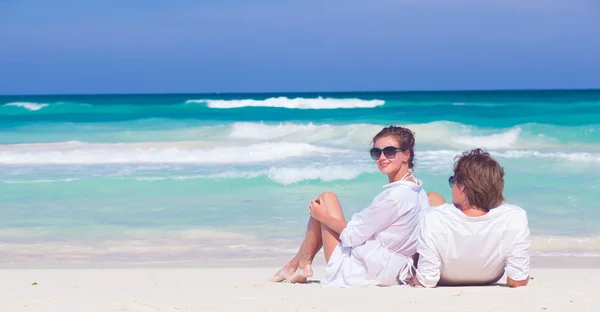 Ritratto di giovane coppia felice in occhiali da sole in abiti bianchi seduta sulla spiaggia tropicale — Foto Stock