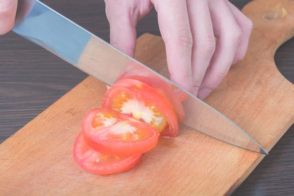 Slice Tomatoes in Half. Red tomato on the chopping Board, in the kitchen. Female Chef cuts one red tomato with a knife on wooden cutting board. The process of cooking, close-up.