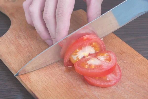 Slice Tomatoes in Half. Red tomato on the chopping Board, in the kitchen. Female Chef cuts one red tomato with a knife on wooden cutting board. The process of cooking, close-up.