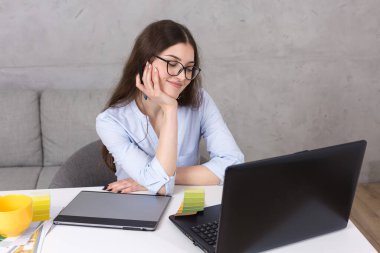 Masada oturan genç bir tasarımcı kız tablete resim çizer. Bilgisayarda iş hesaplamaları yapar.
