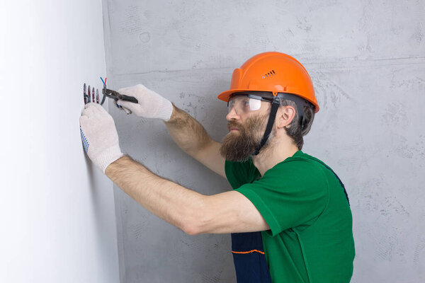 An electrician installs sockets in the apartment. A guy in an orange helmet and overalls makes electrics in the house.