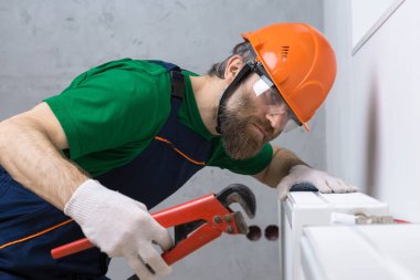 Erkek bir tesisatçı bir apartmanın ısıtma sistemine radyatör yerleştiriyor. Tulumlu ve ingiliz anahtarı olan bir adam..