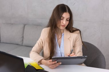 Masada oturan genç bir tasarımcı kız tablete resim çizer. Bilgisayarda iş hesaplamaları yapar.