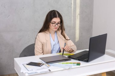 Masada oturan genç bir tasarımcı kız tablete resim çizer. Bilgisayarda iş hesaplamaları yapar.