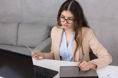 Masada oturan genç bir tasarımcı kız tablete resim çizer. Bilgisayarda iş hesaplamaları yapar.