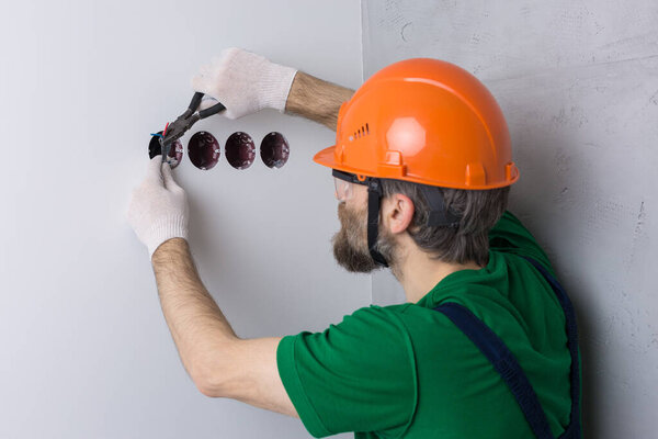 An electrician installs sockets in the apartment. A guy in an orange helmet and overalls makes electrics in the house.