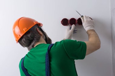 Bir elektrikçi daireye prizler takıyor. Turuncu kasklı ve tulumlu bir adam evde elektrik üretiyor..