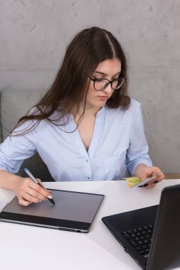 Masada oturan genç bir tasarımcı kız tablete resim çizer. Bilgisayarda iş hesaplamaları yapar.