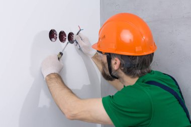 Bir elektrikçi daireye prizler takıyor. Turuncu kasklı ve tulumlu bir adam evde elektrik üretiyor..