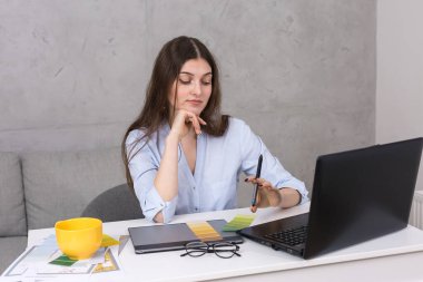 Masada oturan genç bir tasarımcı kız tablete resim çizer. Bilgisayarda iş hesaplamaları yapar.