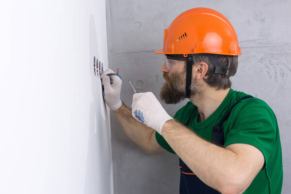 An electrician installs sockets in the apartment. A guy in an orange helmet and overalls makes electrics in the house.
