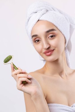 Kalın kaşlı güzel bir kız, beyaz arka planda mükemmel bir ten, kafasında havlu, güzel bir fotoğraf. Kozmetik rulo tutuyor..