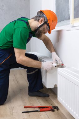 Erkek bir tesisatçı bir apartmanın ısıtma sistemine radyatör yerleştiriyor. Tulumlu ve ingiliz anahtarı olan bir adam..