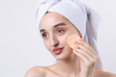 Kalın kaşlı güzel bir kız, beyaz arka planda mükemmel bir ten, kafasında havlu, güzel bir fotoğraf. Kozmetik sünger tutuyor..