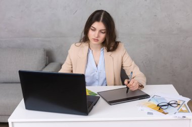 Masada oturan genç bir tasarımcı kız tablete resim çizer. Bilgisayarda iş hesaplamaları yapar.