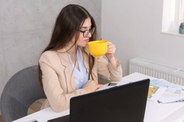 Masada oturan genç bir tasarımcı kız tablete resim çizer. Bilgisayarda iş hesaplamaları yapıyor. Bir kupa kahveden içmek.
