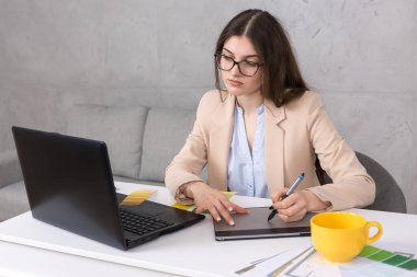 Masada oturan genç bir tasarımcı kız tablete resim çizer. Bilgisayarda iş hesaplamaları yapar.