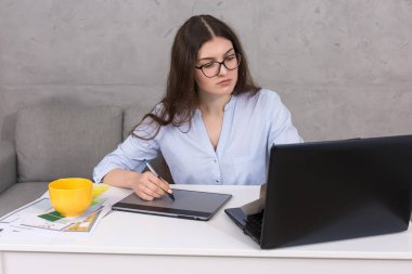 Masada oturan genç bir tasarımcı kız tablete resim çizer. Bilgisayarda iş hesaplamaları yapar.