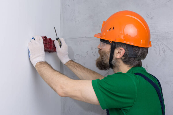 An electrician installs sockets in the apartment. A guy in an orange helmet and overalls makes electrics in the house.