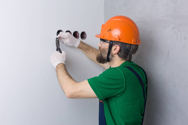 An electrician installs sockets in the apartment. A guy in an orange helmet and overalls makes electrics in the house.