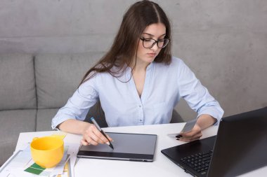 Masada oturan genç bir tasarımcı kız tablete resim çizer. Bilgisayarda iş hesaplamaları yapar.